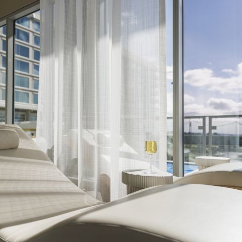 A sunlit room features a modern lounge chair, sheer curtains, and floor-to-ceiling windows with a view of a balcony and the sky.