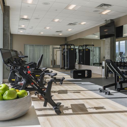 A modern gym with exercise bikes, weights, and workout equipment. A bowl of green apples is on a table in the foreground.