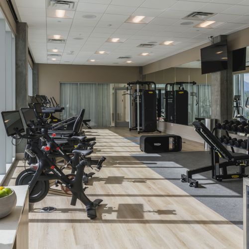 A modern gym with exercise bikes, weightlifting equipment, a TV, and towels; windows let in natural light.