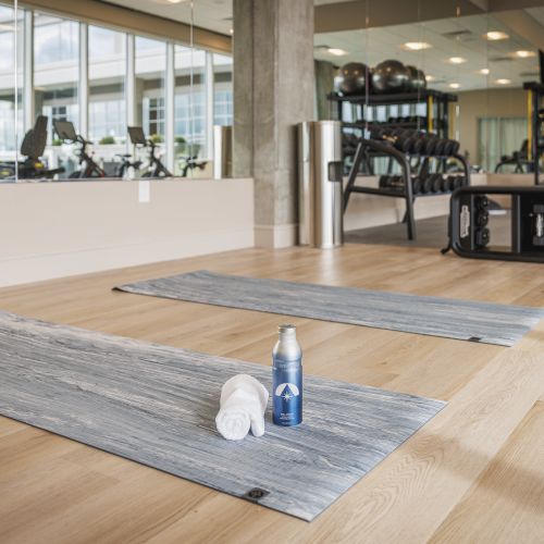 The image shows a gym with exercise mats, a towel, and a water bottle on a wooden floor, with fitness equipment in the background.