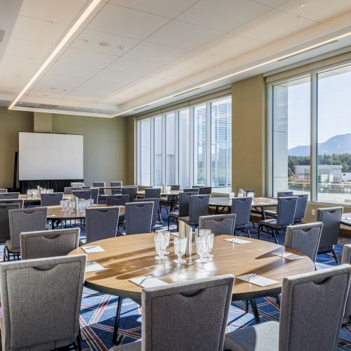 A conference room with arranged tables, chairs, a projector screen, and large windows offering a view of the outdoors.