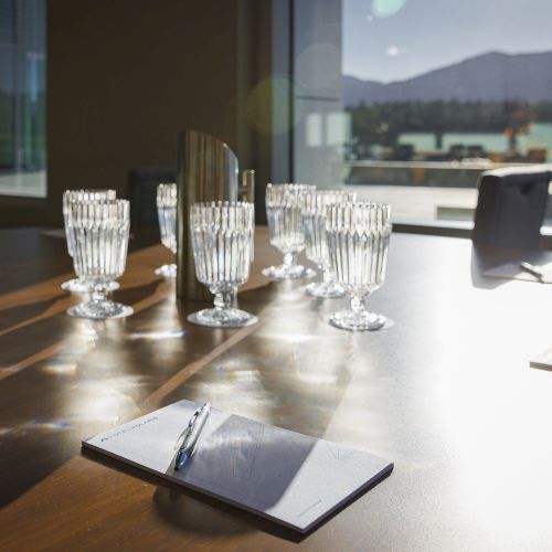 The image shows a conference table with notepads, pens, and glass tumblers, set in a bright room with a view of mountains outside.