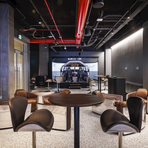 A modern lounge with round tables and unique chairs in the foreground, and a flight simulator setup in the background.