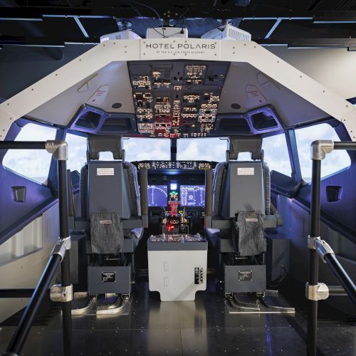 The image shows the interior of a flight simulator cockpit with two pilot seats and control panels, set in a simulated environment.