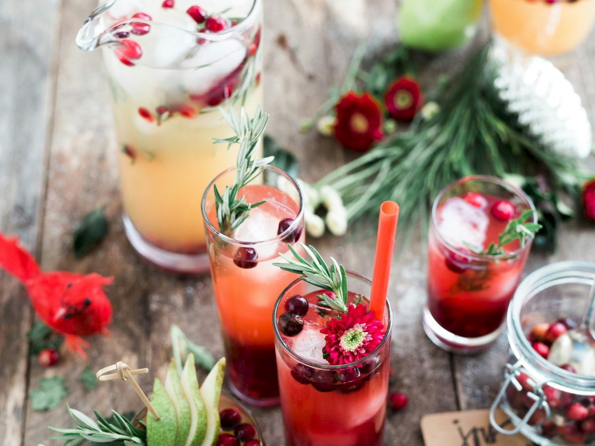 A rustic setup with colorful drinks garnished with fruit and herbs on a wooden table, surrounded by flowers and festive decorations.