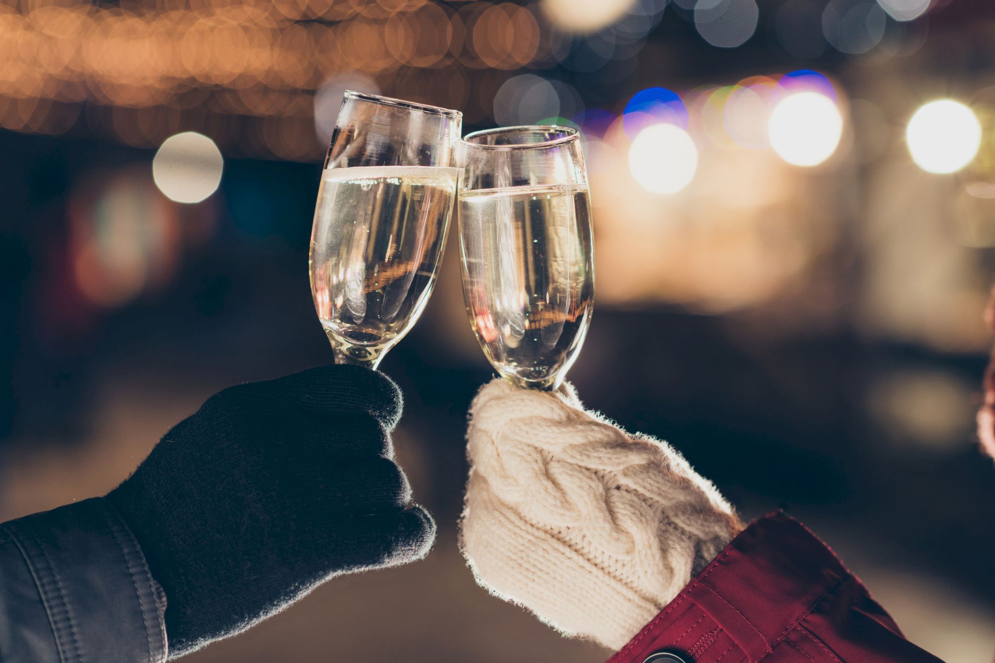 Two people clinking champagne glasses while wearing gloves, against a blurred festive background.