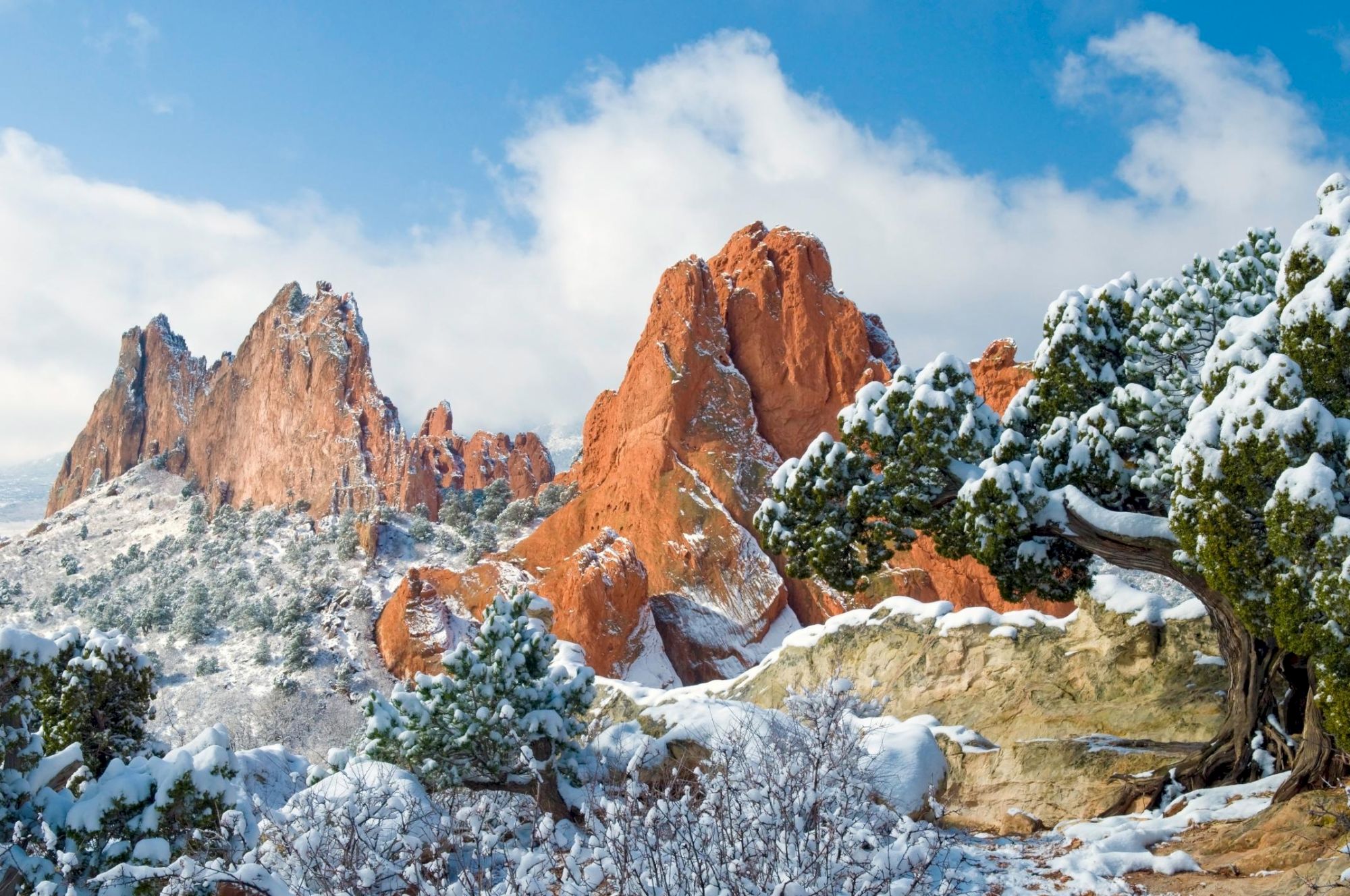 Snow-covered red rock formations and trees under a blue sky with clouds.