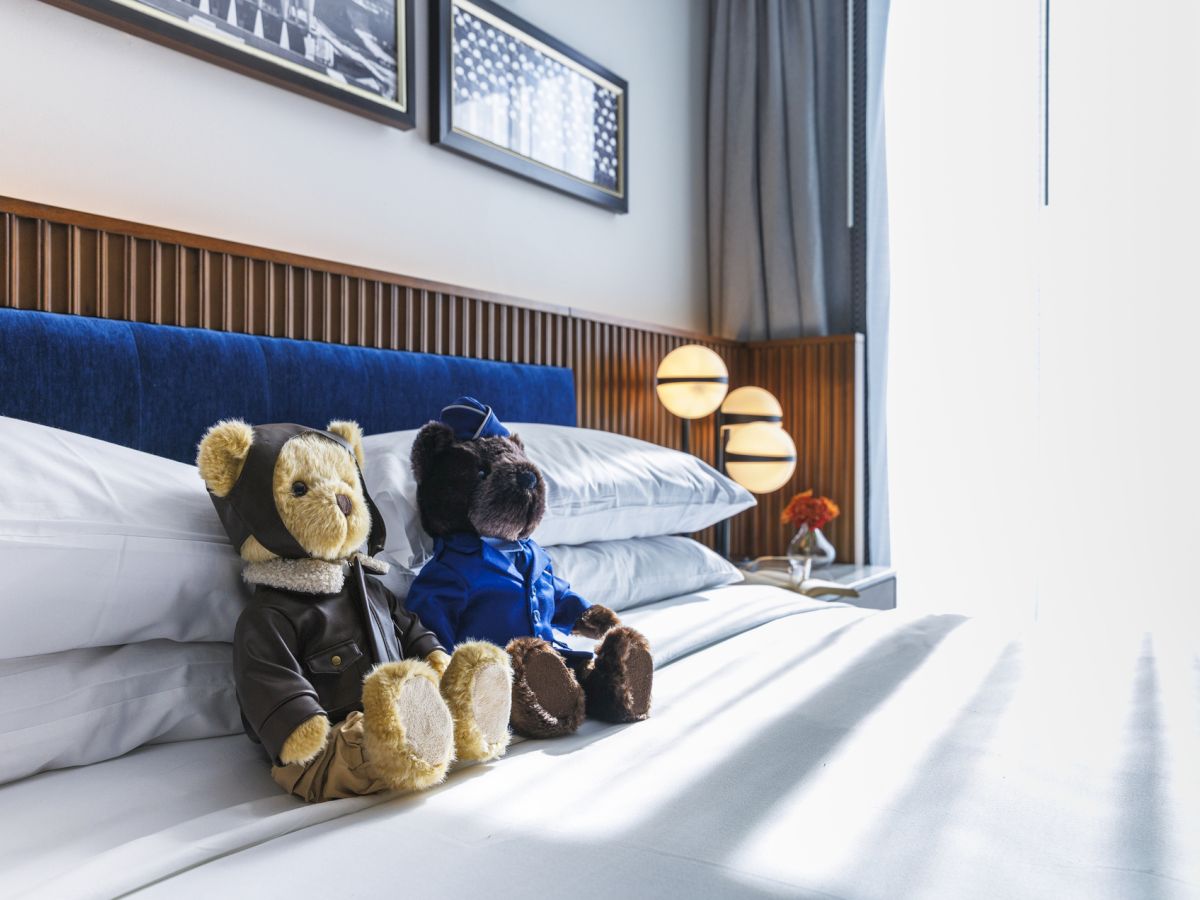 Two teddy bears in outfits sit on a neatly made bed with white linens, beside a wooden headboard and modern lamps.