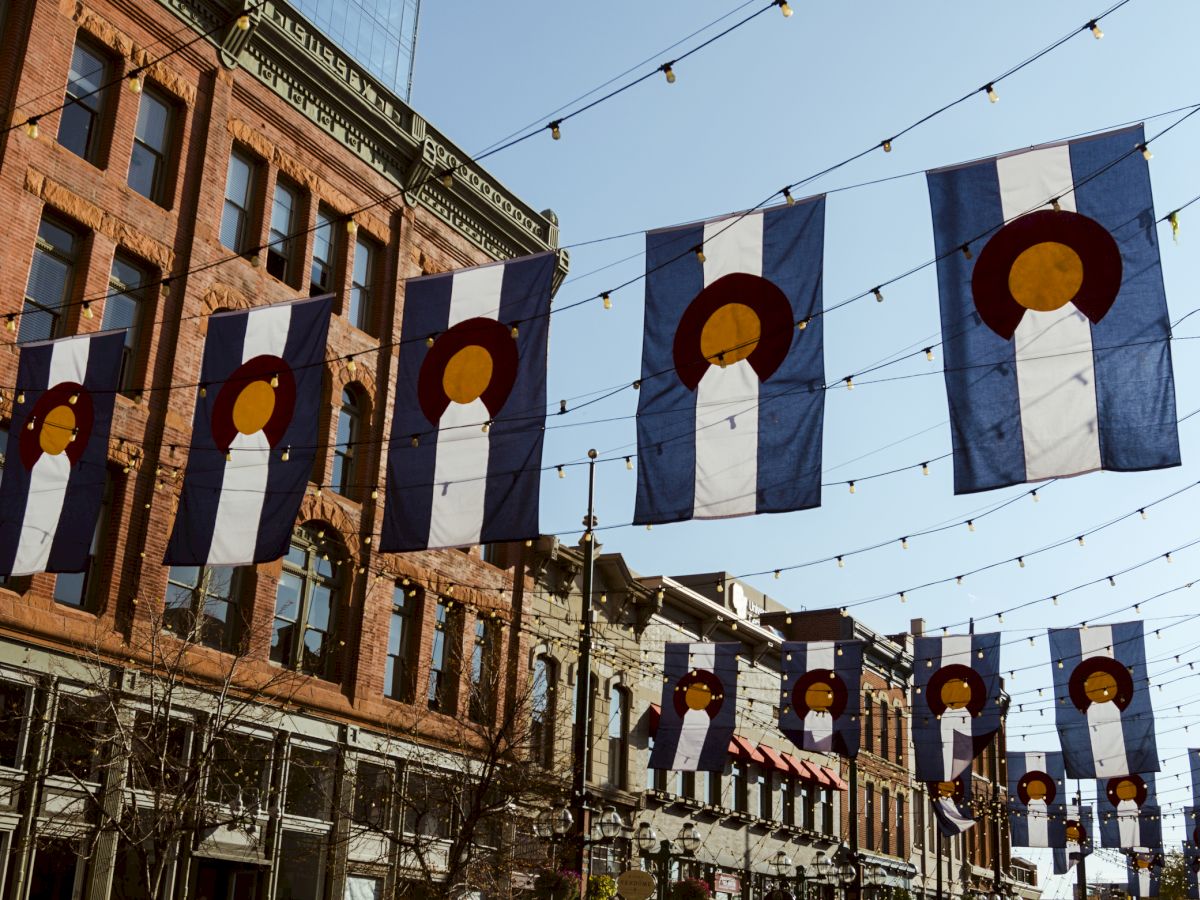 The image shows a street lined with Colorado flags hanging over a row of brick buildings, decorated with string lights under a clear sky.