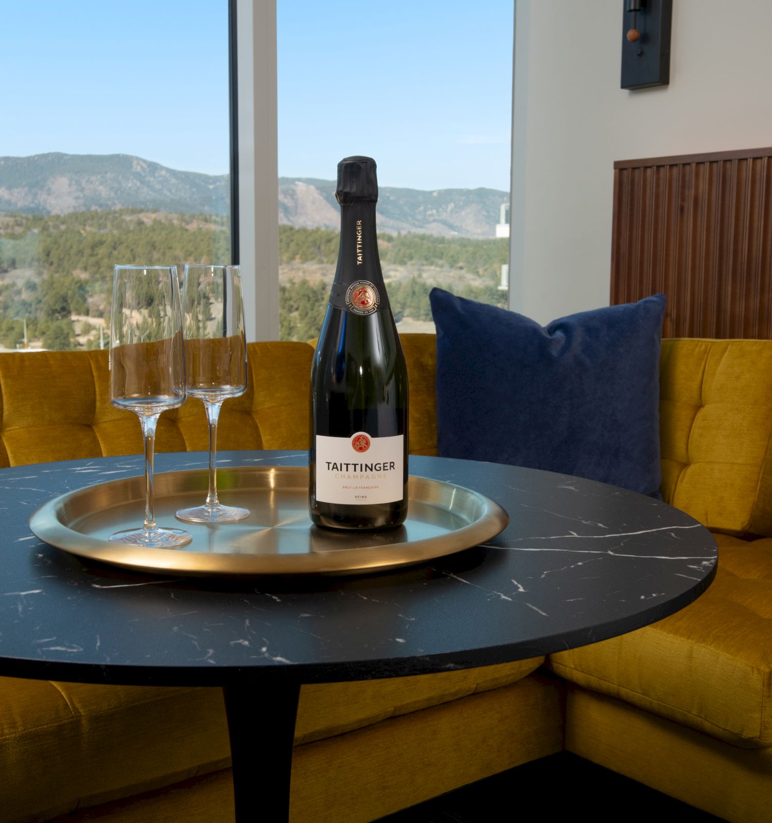 A bottle and two glasses sit on a table with a mountain view in the background, complemented by yellow seating.