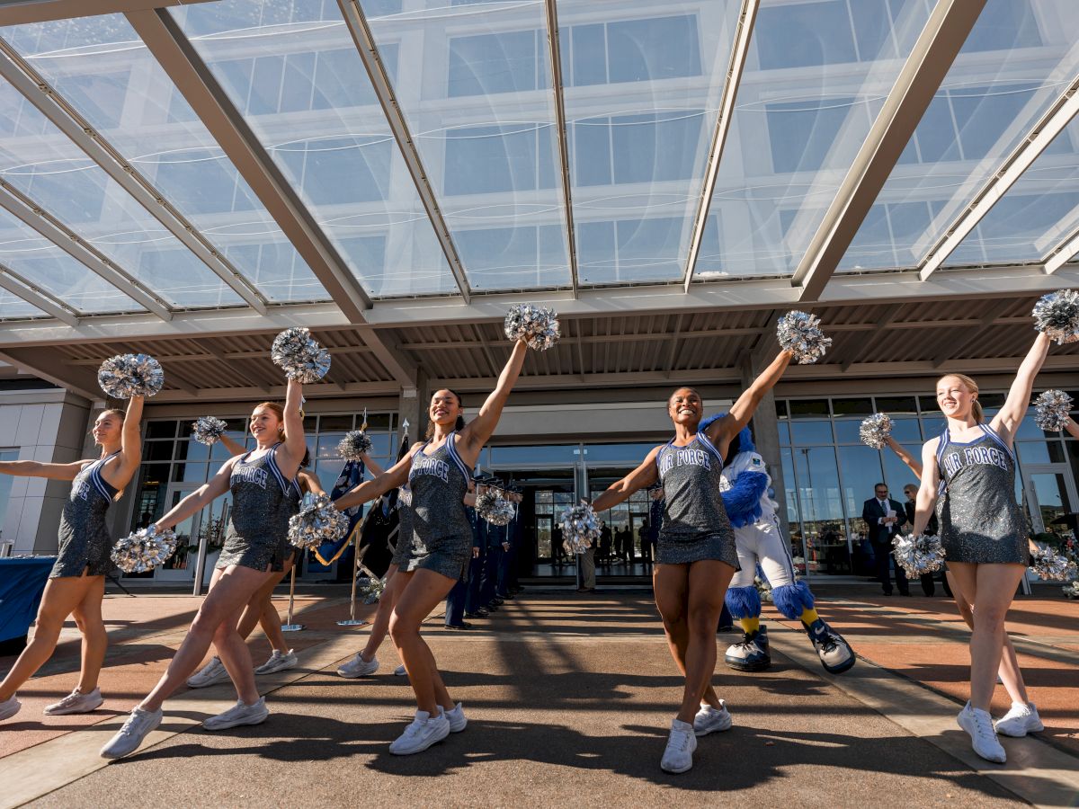 A group of cheerleaders performs a routine with pom-poms in front of a building with a glass canopy.