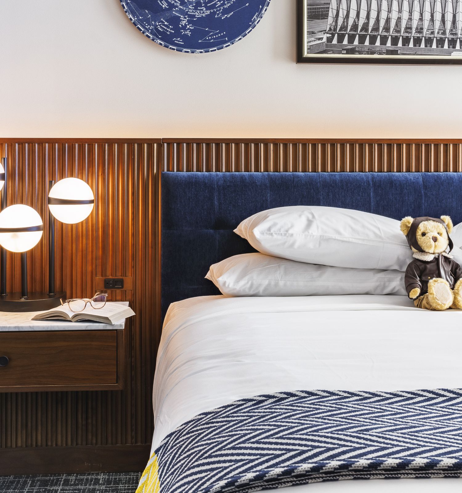 A bed with two teddy bears on it, a side table with a lamp and book, and framed pictures on the wall.