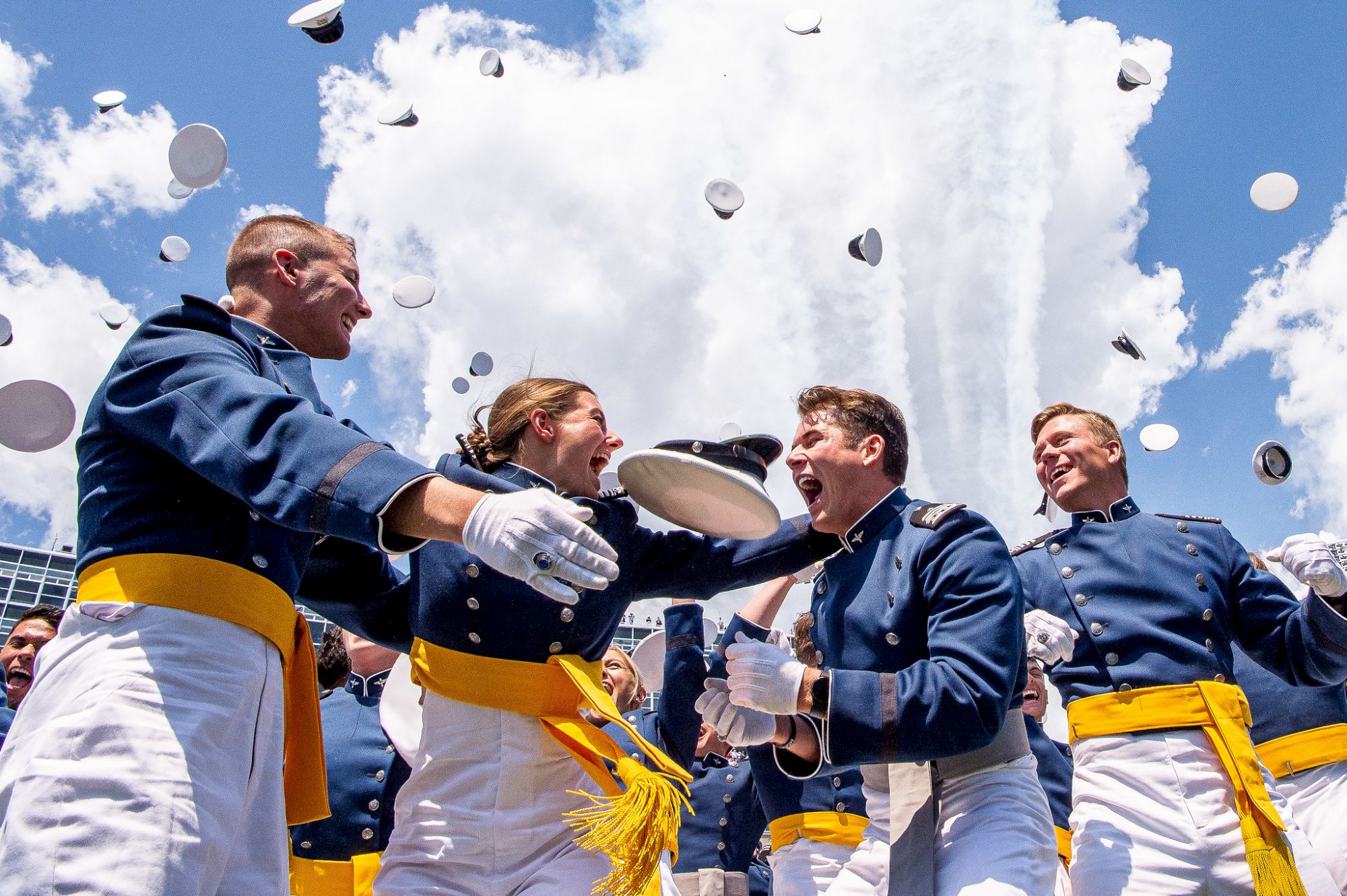 Cadets in uniform celebrate by tossing hats in the air against a blue sky, with white clouds and a plane's trail in the background.