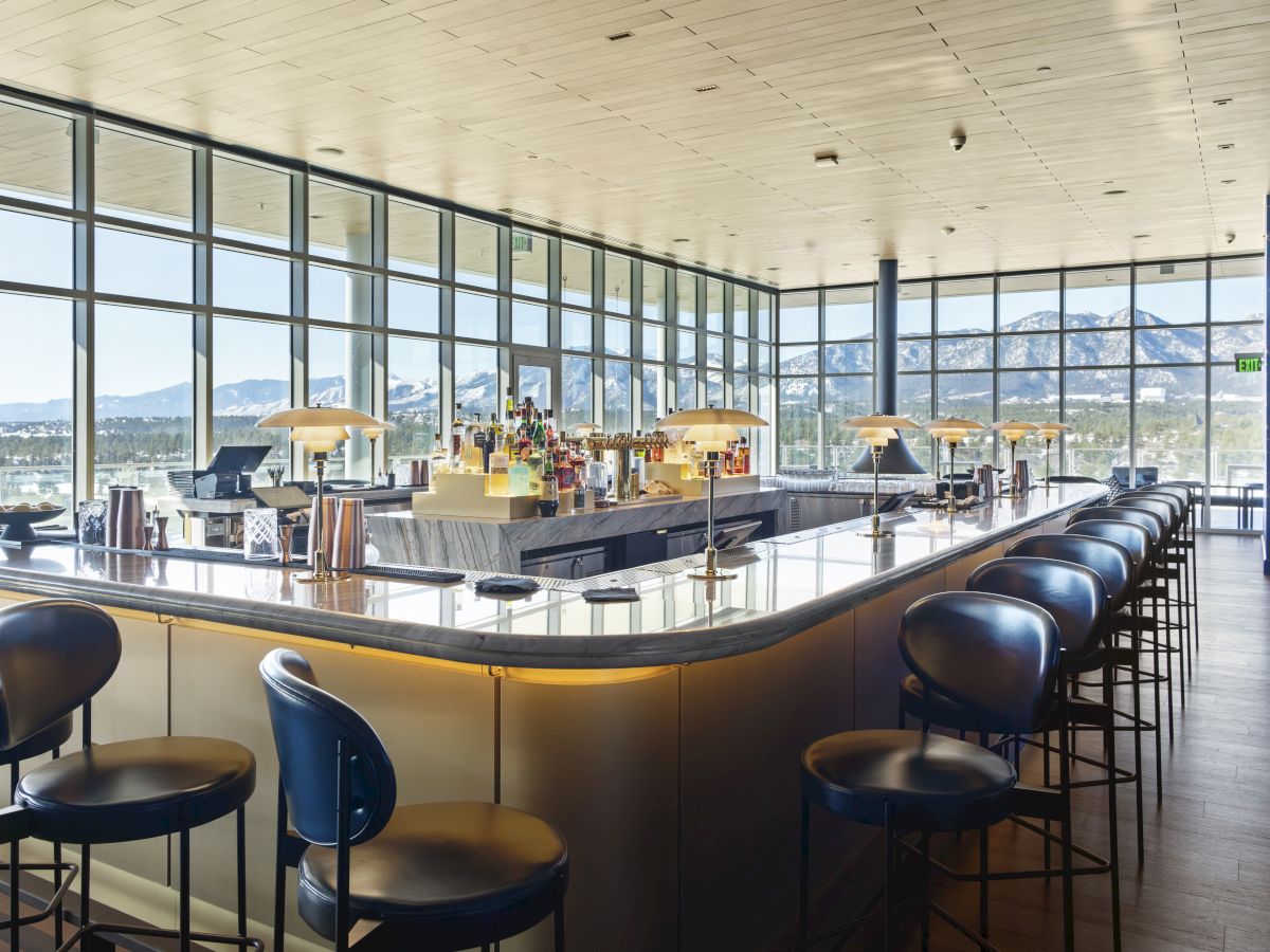 The image shows a modern bar with stools, a mirrored counter, and large windows offering a scenic mountain view in the background.