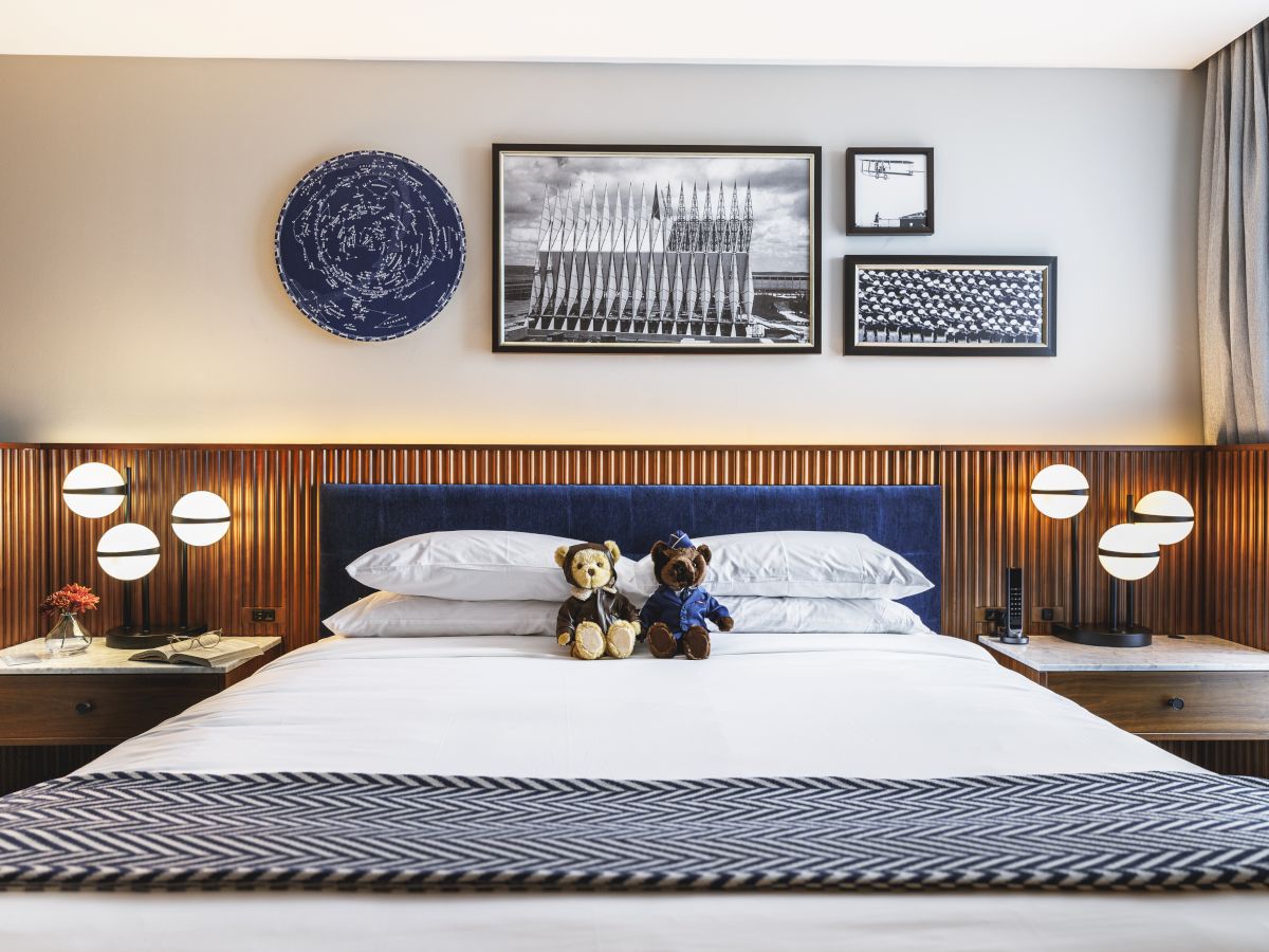 Cozy bedroom with a double bed, bears, and geometric lamps. Wall art decorates the neutral wall. Gray curtains frame the scene.