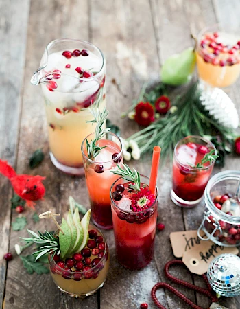 A variety of colorful drinks garnished with fruits and herbs on a wooden table, surrounded by flowers and festive decorations.