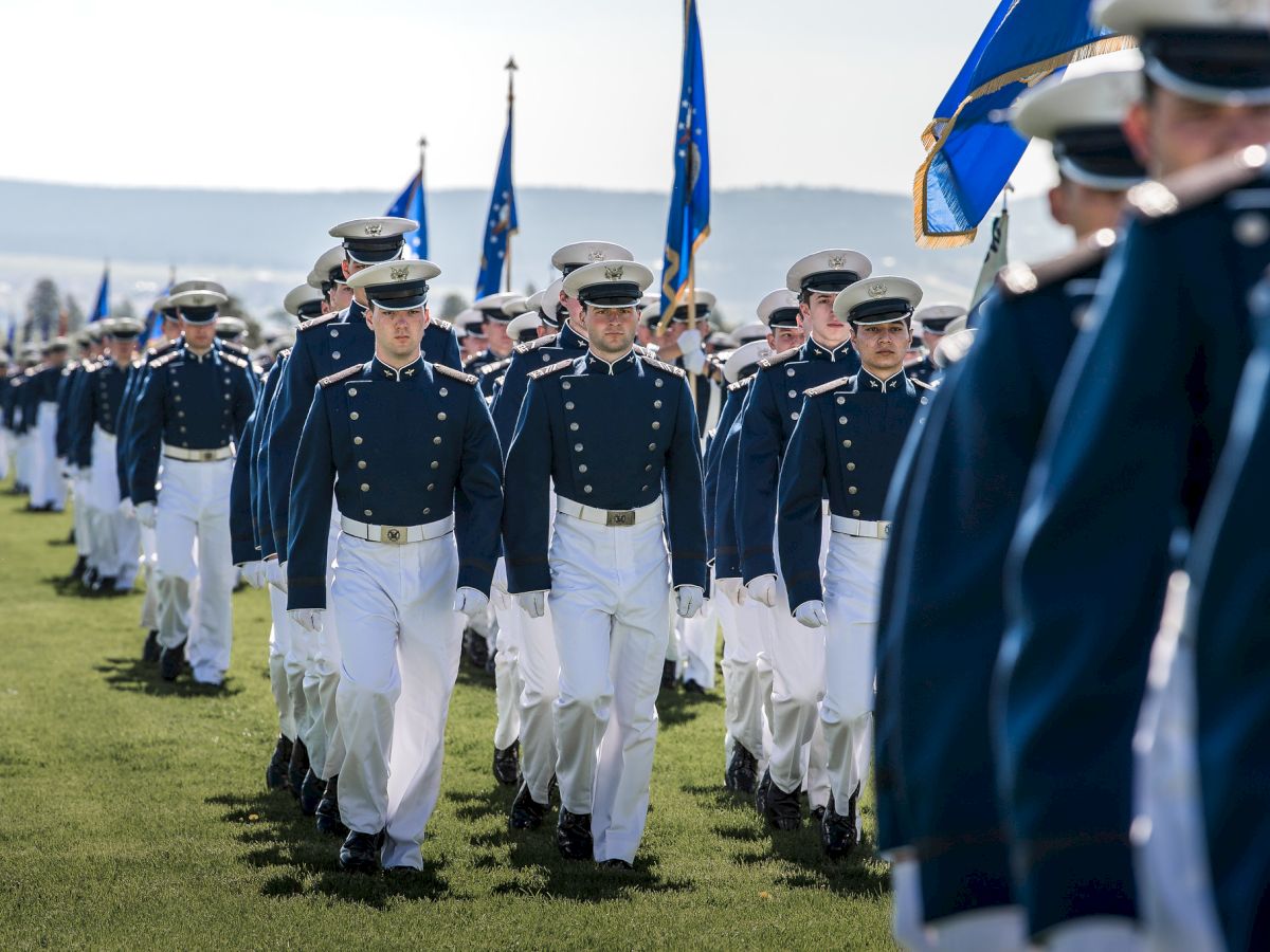 A group of people in uniform marching in formation, holding flags, on a grassy field.