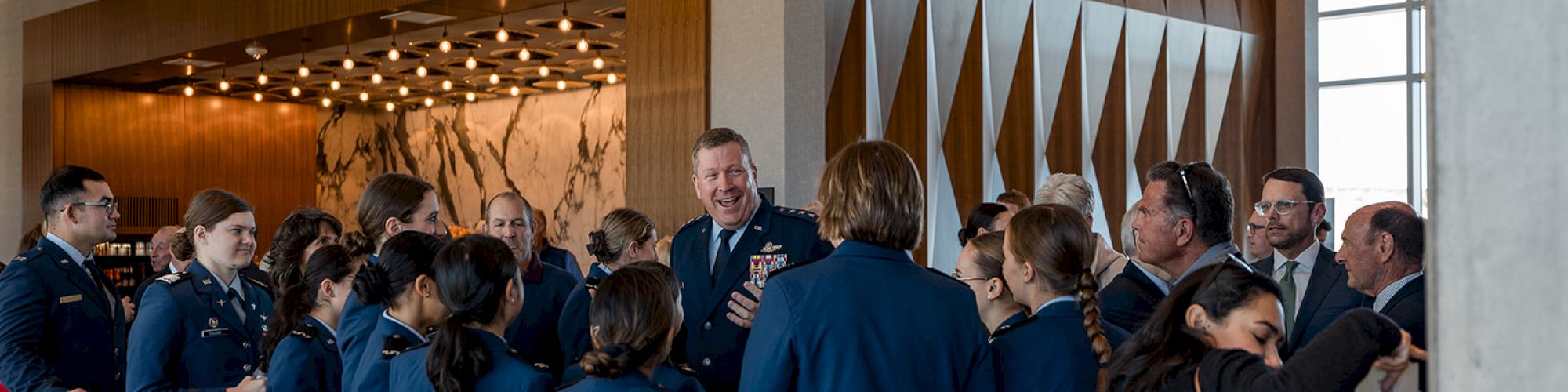 A group of people in uniform are gathered in an indoor setting with wood paneling and artwork in the background.