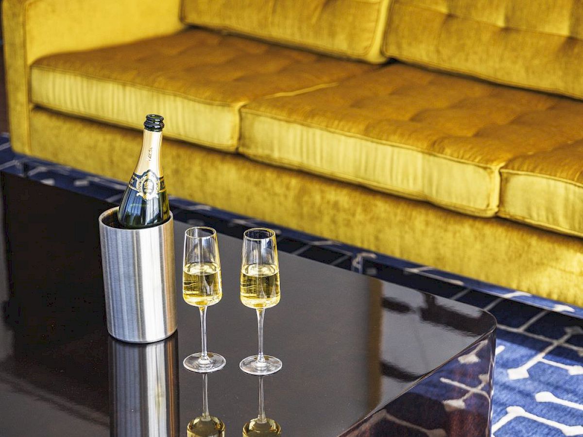 A yellow couch with two glasses of champagne and a bottle in an ice bucket on a glass table in front of it.