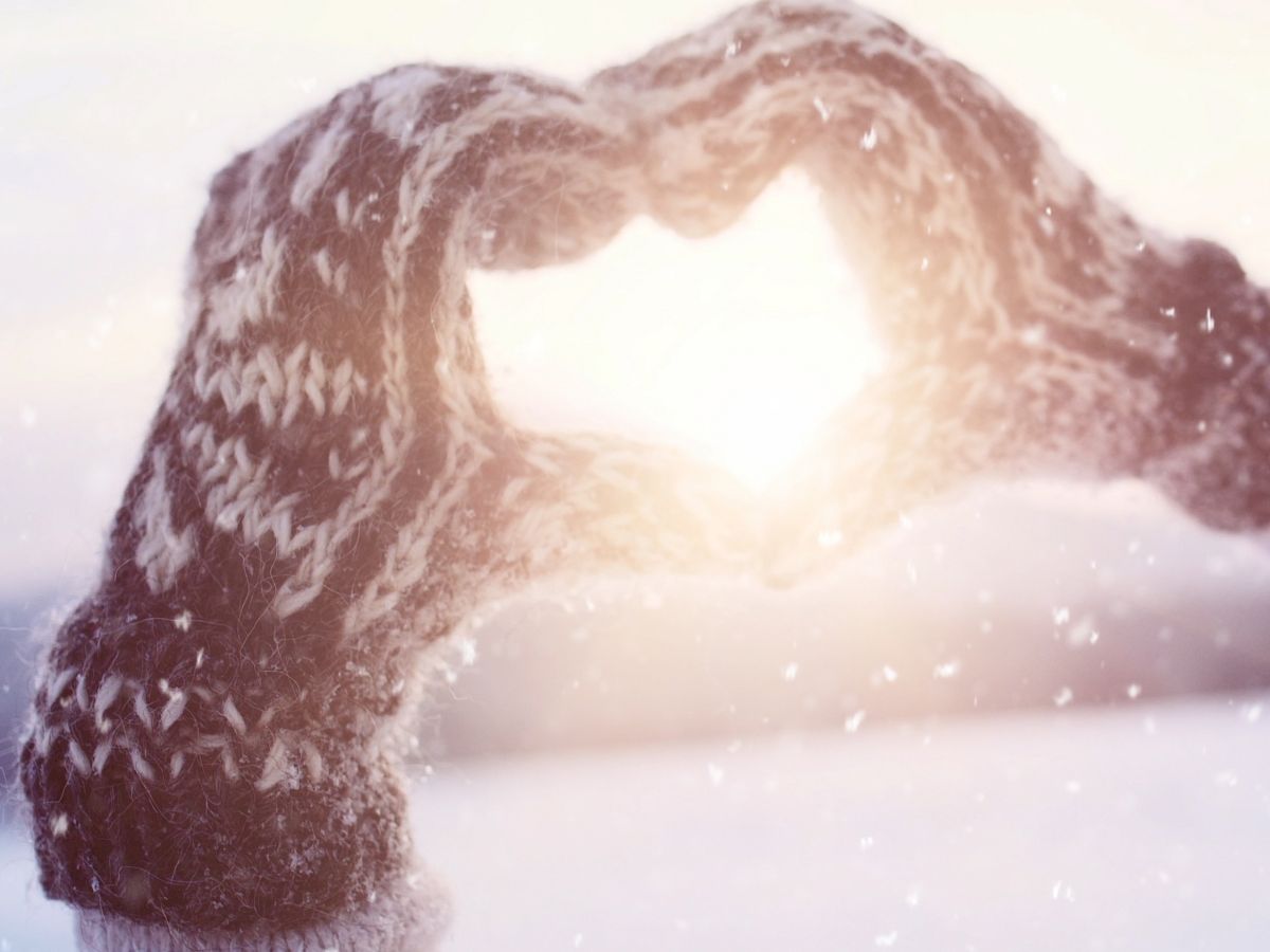 Warm knitted gloves form a heart shape against a snowy backdrop, with sunlight shining through in a winter scene.