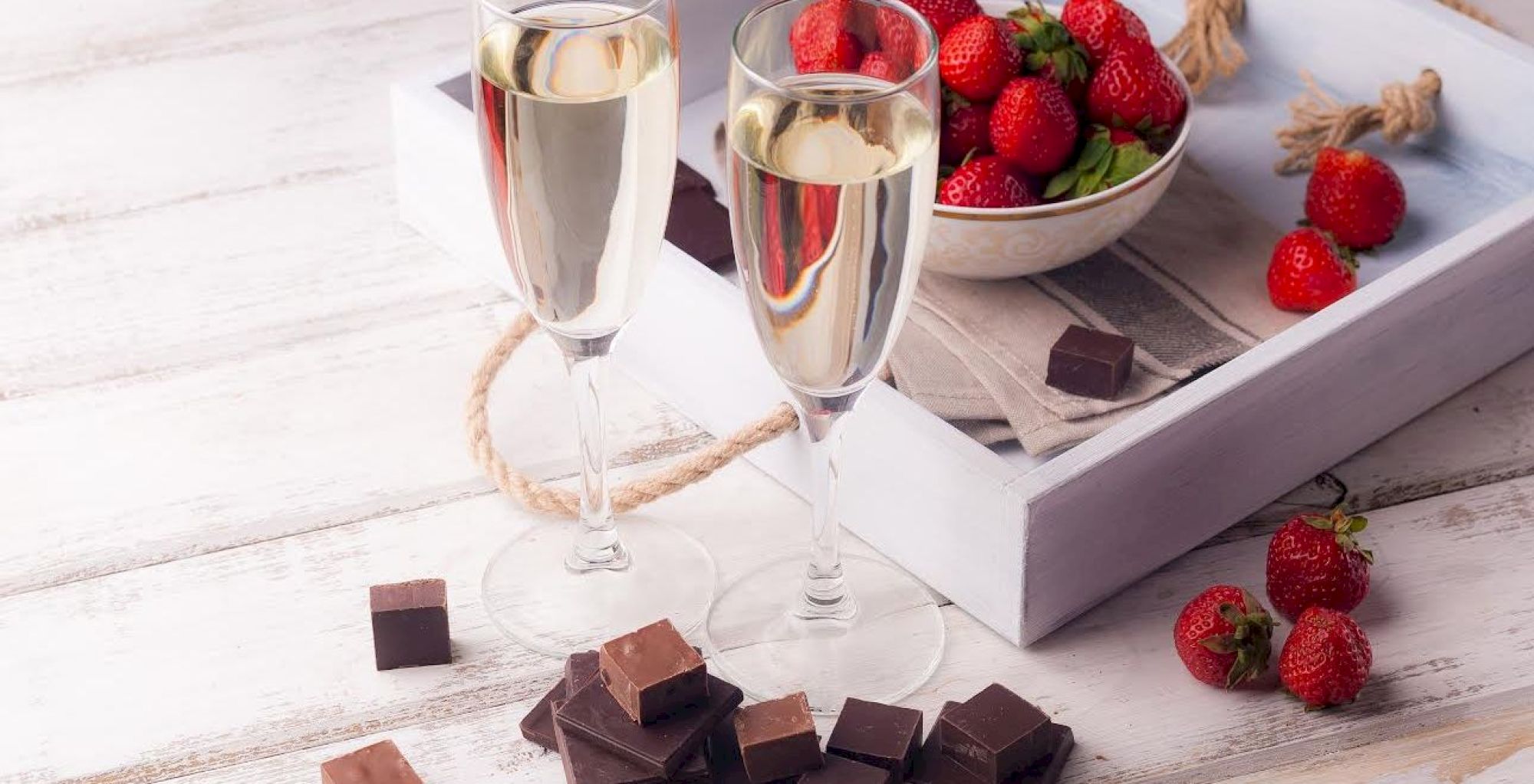 The image shows two glasses of champagne, a bowl of strawberries, and pieces of chocolate on a white wooden surface.