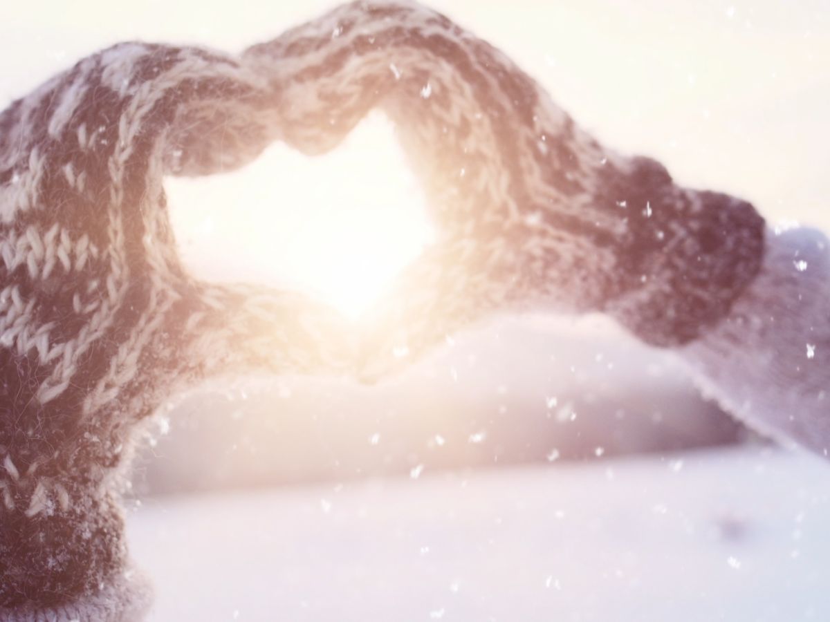 Two hands in knitted gloves form a heart shape in front of the sun, with snow falling around, creating a cozy winter scene.