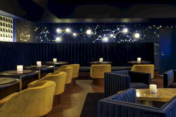 A stylish lounge with dark velvet seating, round tables, yellow chairs, and starry wall decorations, lit by soft ambient lights.