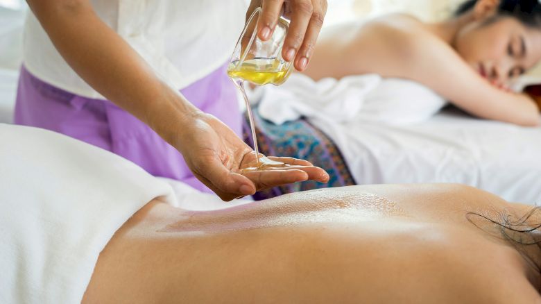 A person receiving a massage with oil being poured on their back, while another person relaxes in the background, wrapped in a towel.