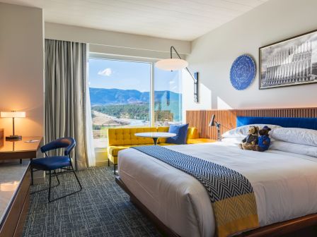 A cozy hotel room with a plush bed, yellow sofa, and scenic window view. Decor includes a mountain photo and a teddy bear on the bed.