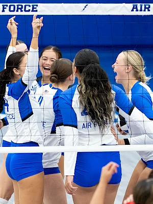 A volleyball team in blue and white jerseys celebrates on the court as players in red jerseys walk by in the foreground.