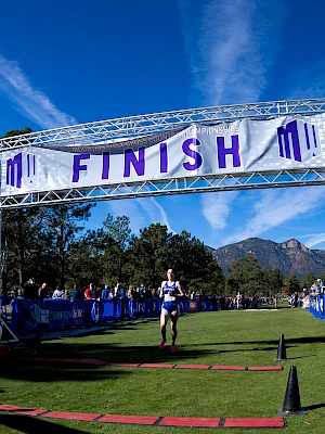 A runner crosses the finish line on a grassy course, surrounded by spectators and blue barriers under a clear sky with mountain views.