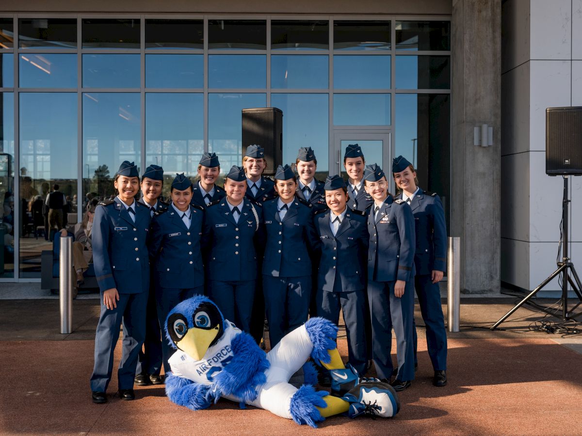 A group in uniform poses with a blue bird mascot lying in front. They stand in front of a building with glass doors.