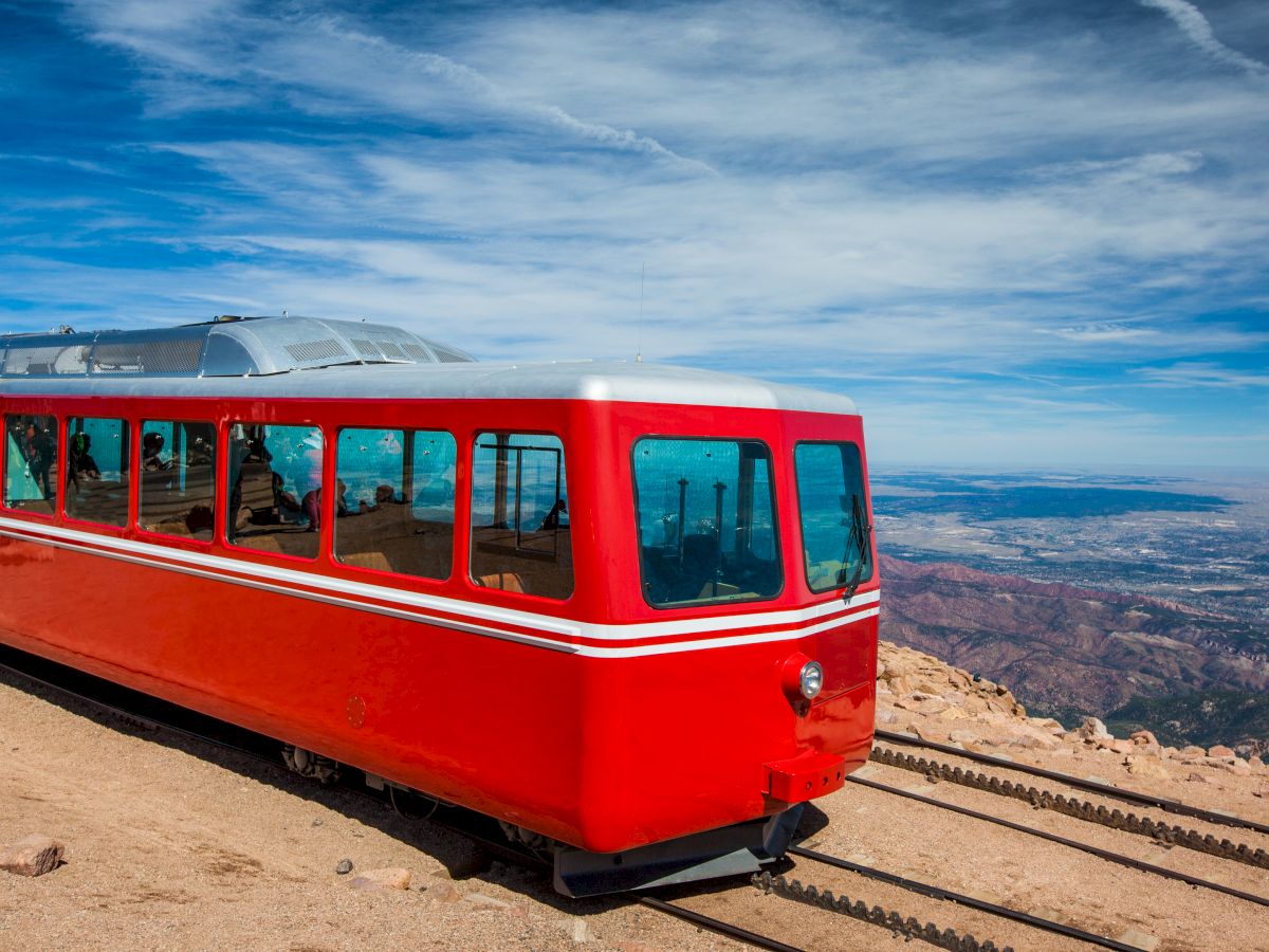 A red train is on tracks near a mountaintop, with a vast landscape and cloudy blue sky in the background.