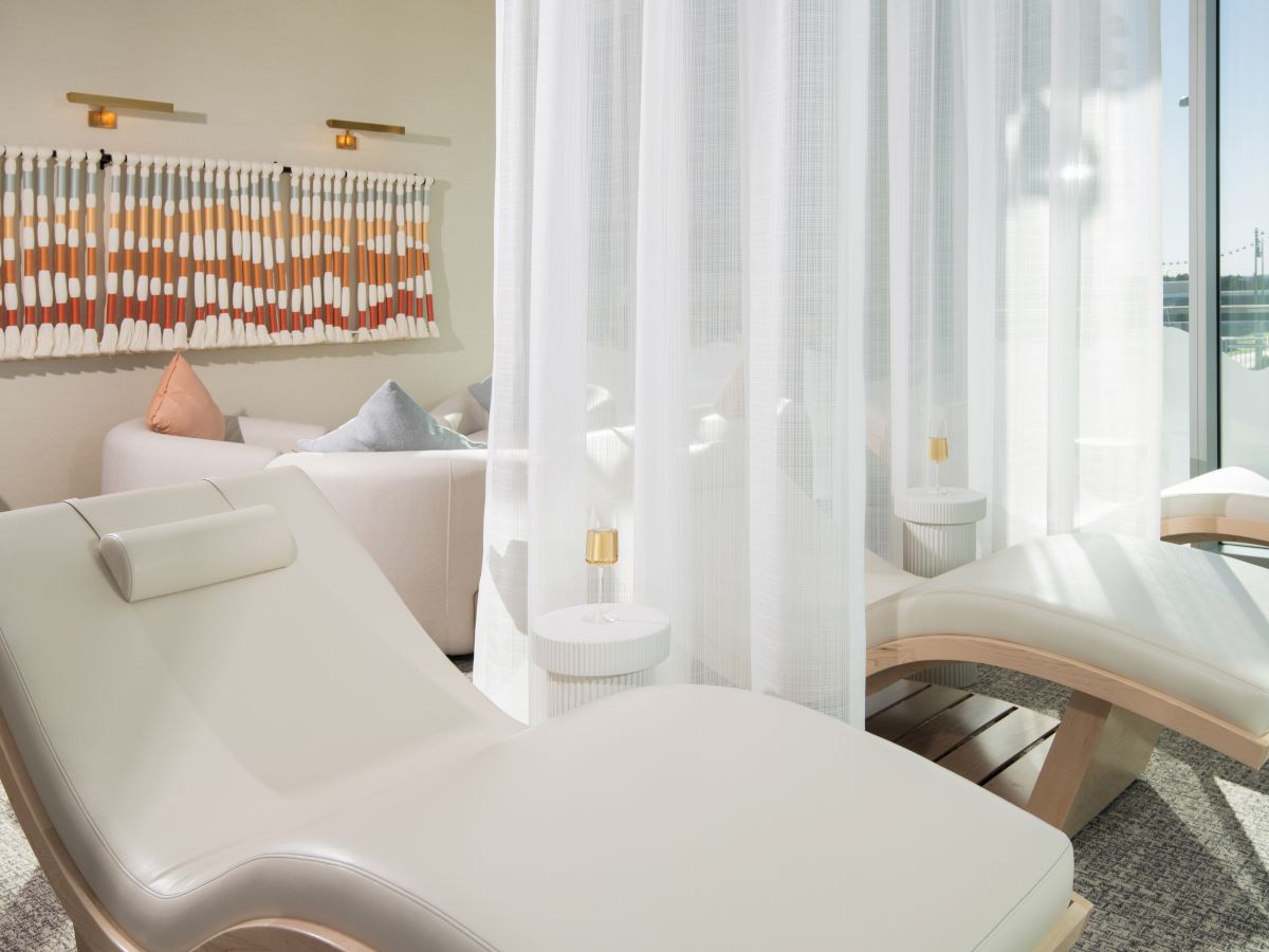The image shows a bright, modern lounge area with white loungers, sheer curtains, and decorative pillows on a couch.