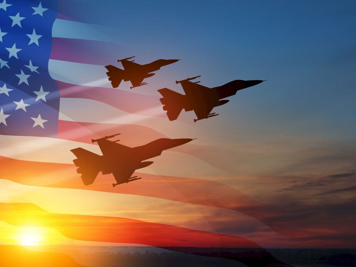Silhouettes of three jet fighters are flying with the American flag in the background against a sunset.
