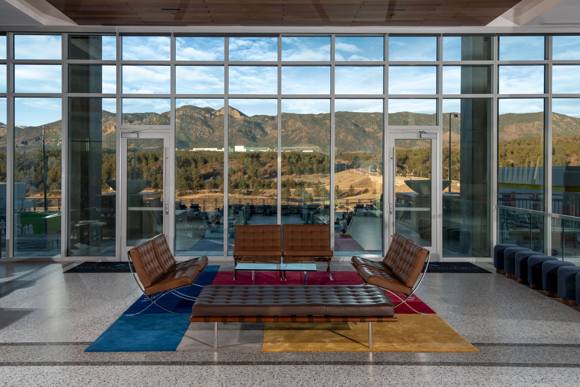 A modern lounge area with chairs and tables, large windows showcasing a scenic view of mountains and trees, and a colorful rug.