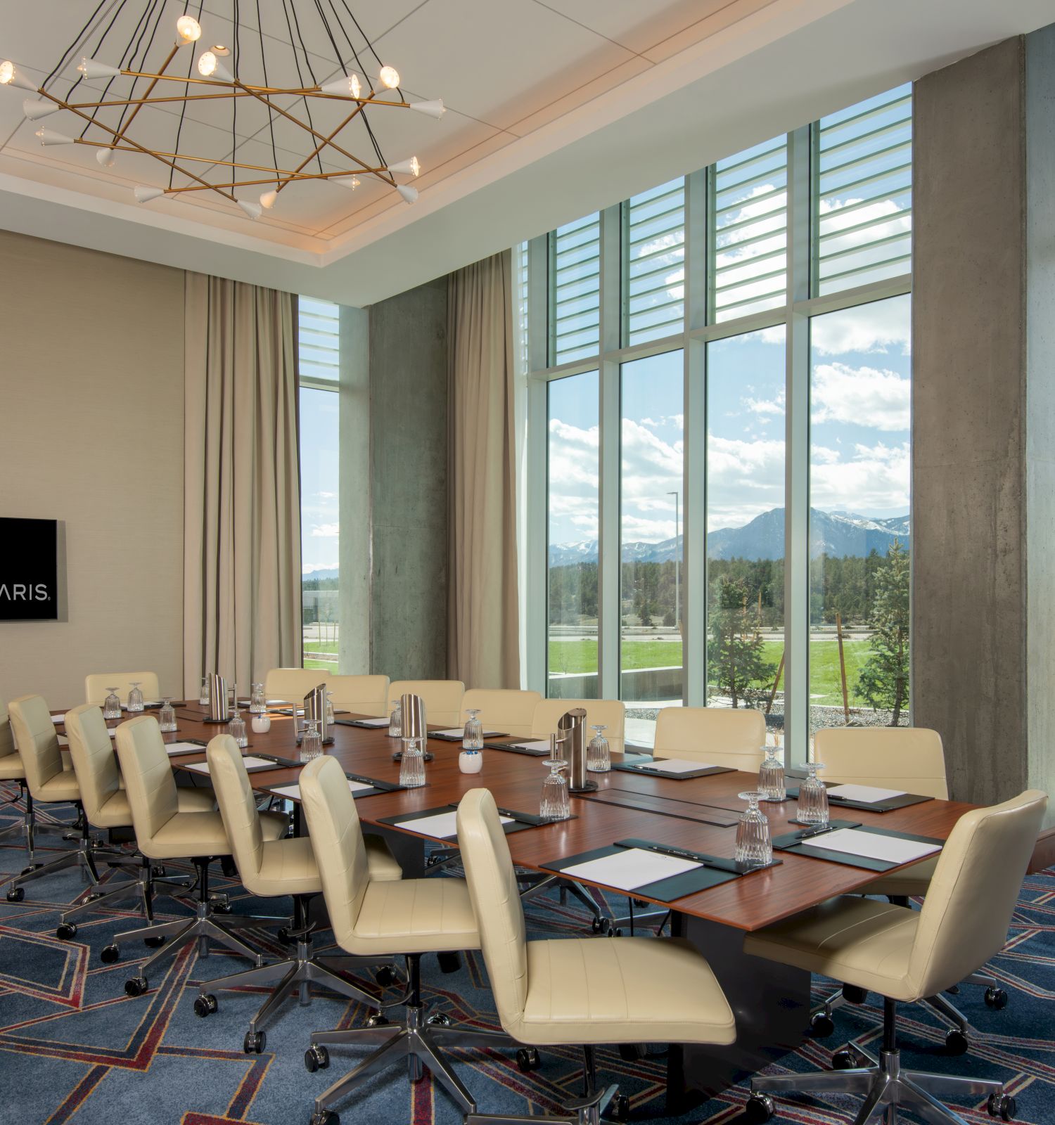 The image shows a modern conference room with a long table, chairs, a screen, and large windows offering a scenic view of mountains and fields.