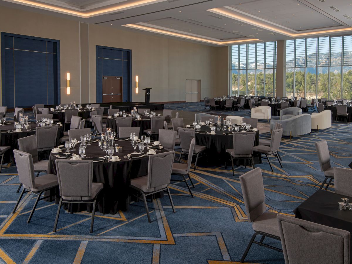 The image shows a spacious banquet hall with round tables set up for an event, featuring large windows and a patterned carpet.