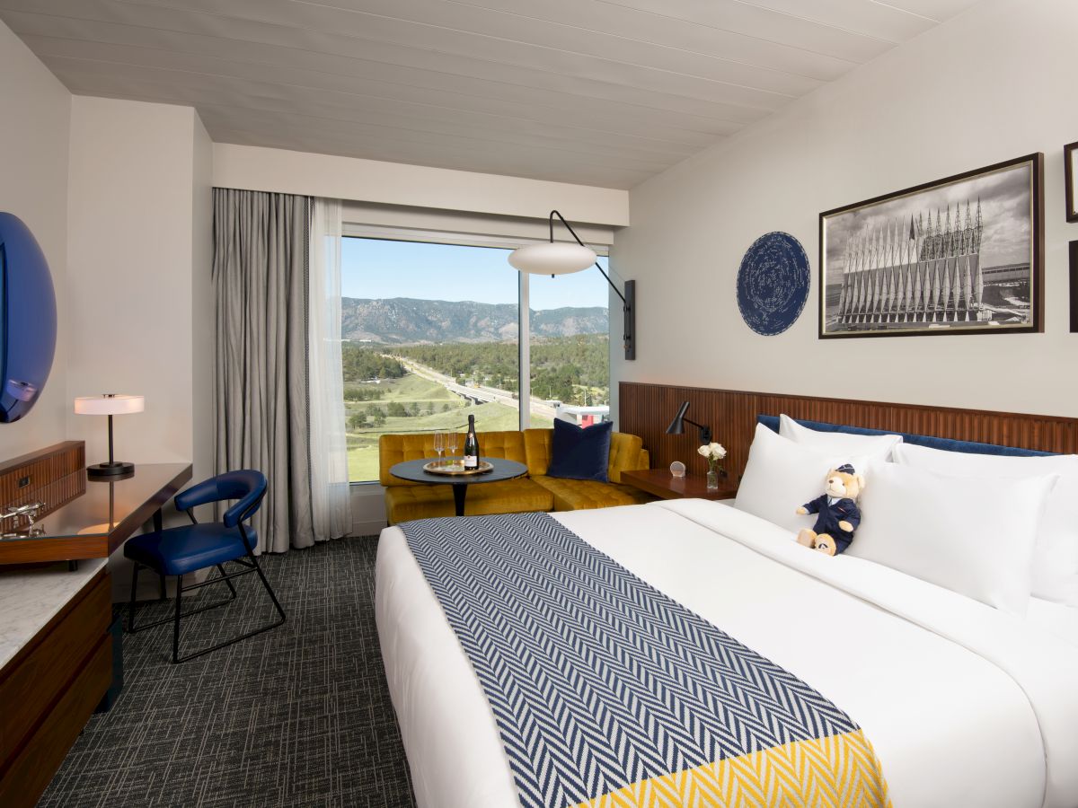 A hotel room with a large bed, desk, chair, and window view of mountains. There are decorative elements and framed art on the walls.