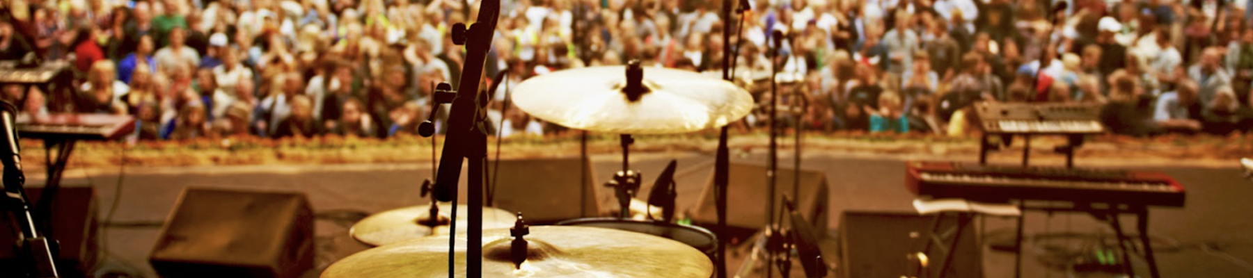 A drum set on stage is set up, facing a large crowd gathered in an outdoor venue for a concert.