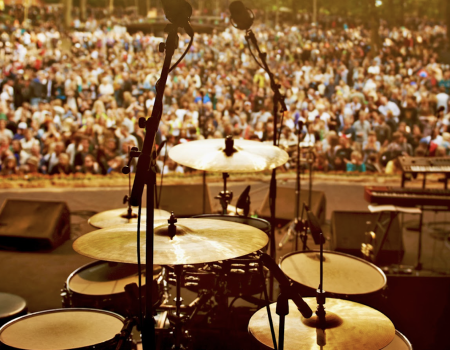 A drum set on stage is set up, facing a large crowd gathered in an outdoor venue for a concert.