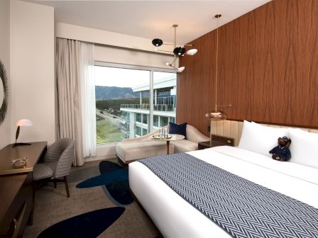 A modern hotel room with a large bed, a window view, a desk, and a cozy seating area with a stuffed bear sitting on the bed.