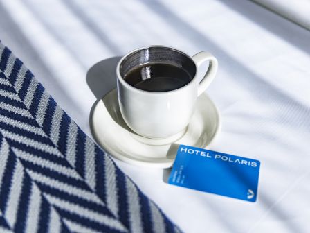 A cup of coffee sits on a saucer next to a blue "Hotel Polaris" card on a bed with a striped blanket.