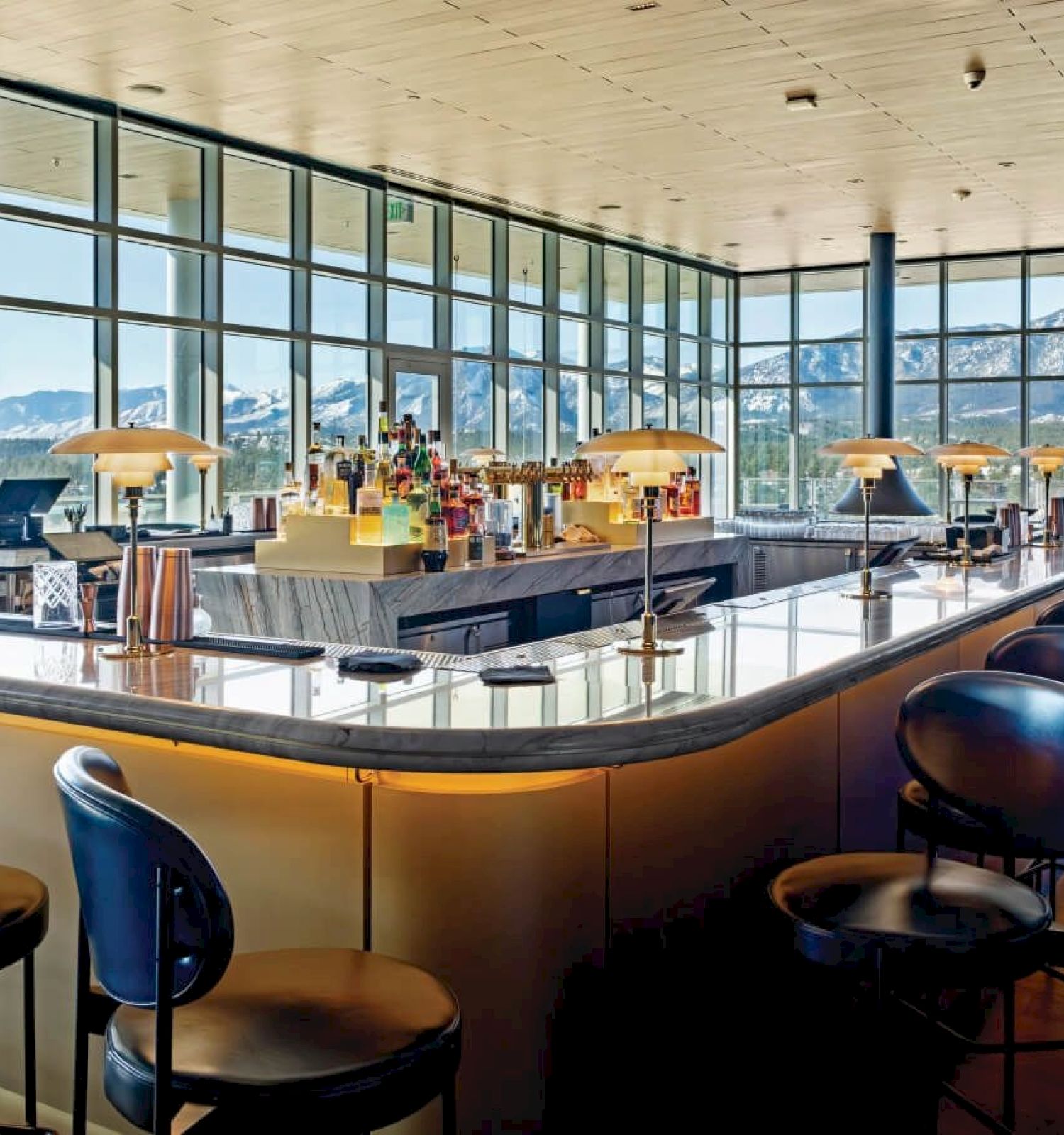 Spacious bar with high chairs, a sleek counter, and panoramic mountain views through large windows under a wooden ceiling.