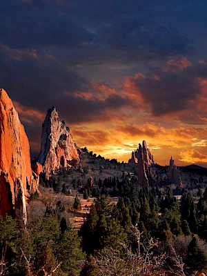 A dramatic sunset illuminates red rock formations and evergreen trees in a stunning landscape scene under a richly colored sky.