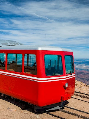 The image shows a red train on a mountain track against a scenic background, with a vast landscape and blue sky in the distance.