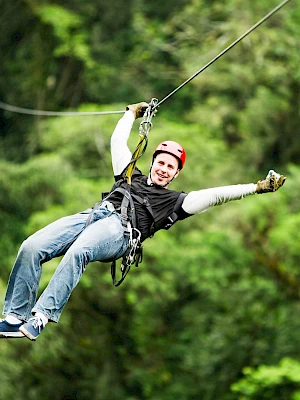 A person is zip-lining through a lush green forest, smiling and wearing casual clothes with safety gear.