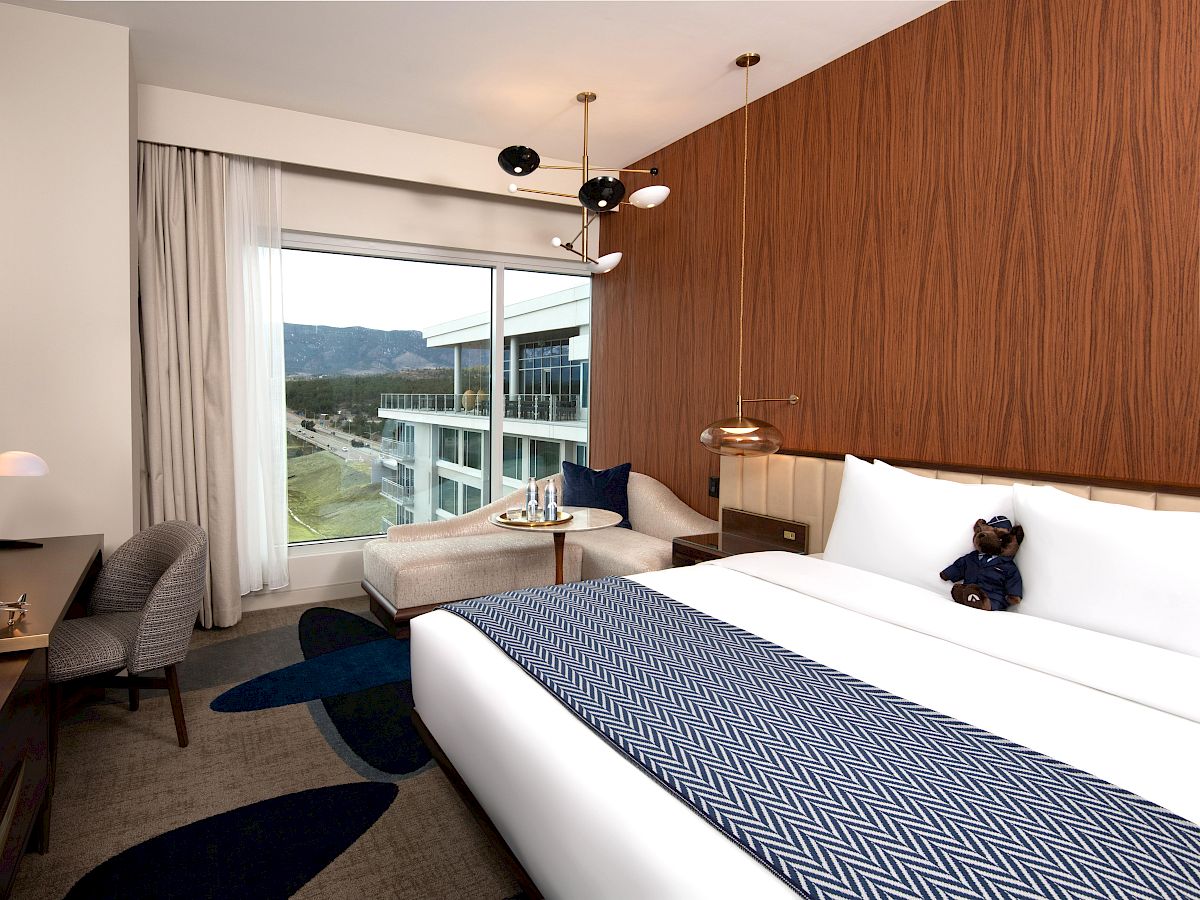 A modern hotel room with a large bed, stylish decor, a window view, and a small sitting area with a teddy bear on the bed.