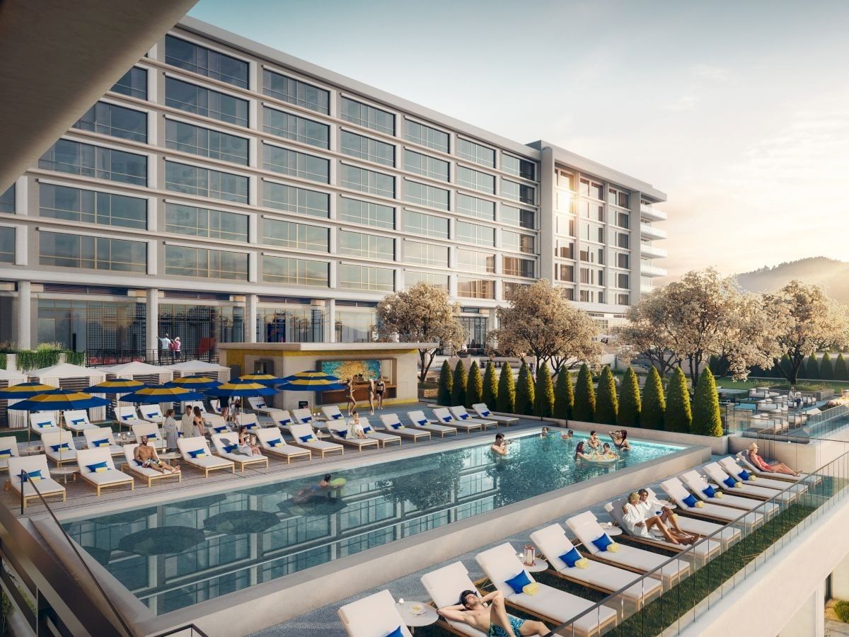 A modern hotel with a spacious outdoor pool area, surrounded by lounge chairs and guests relaxing, set against a mountainous backdrop.