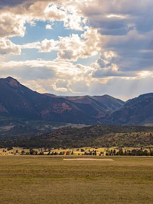 A scenic landscape with mountains under a partly cloudy sky, sunlight streaming through, and a field in the foreground.